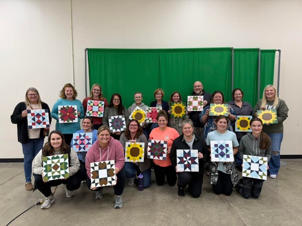 Group of people indoors holding colorful quilt squares in front of green curtains