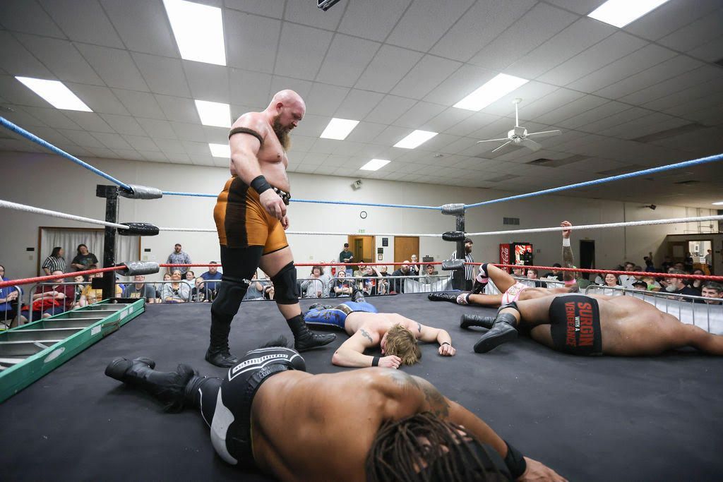 Wrestlers lying on the mat while one stands over them in a gym ring, with spectators in the background