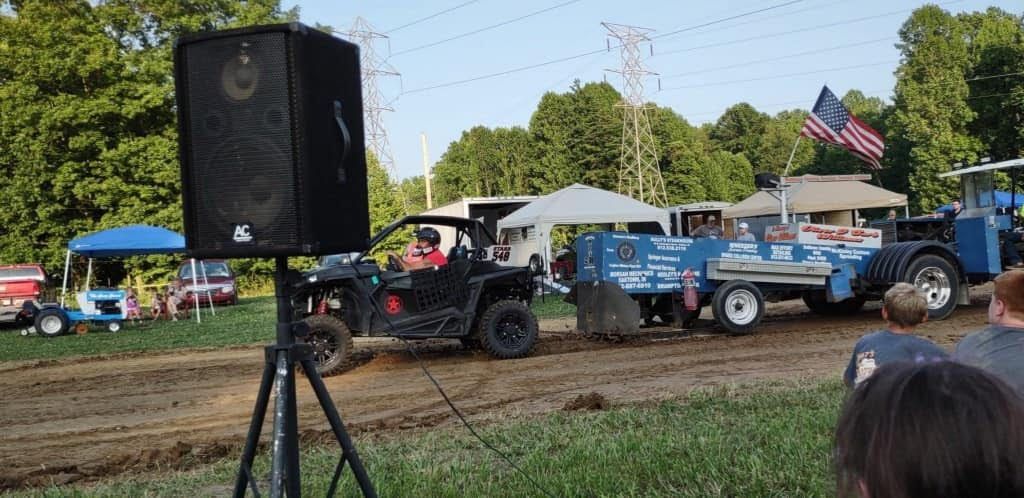 Outdoor tractor pull with a blue truck, black loudspeaker, and spectators near the track