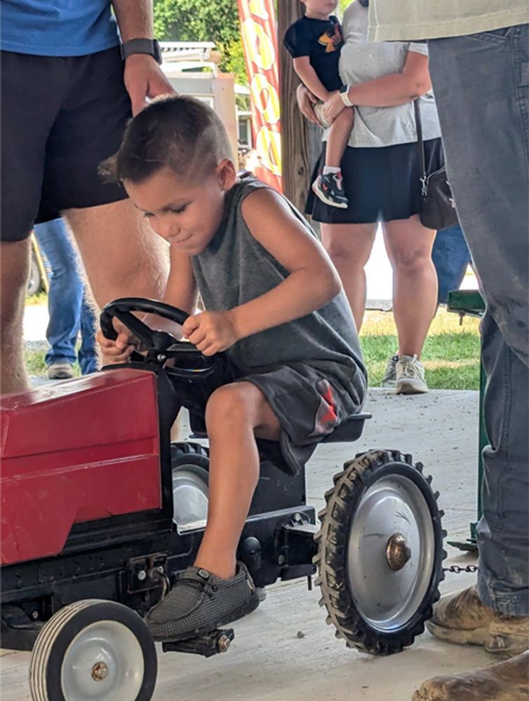 Young child steering a small red ride-on tractor at an outdoor event, surrounded by adults.