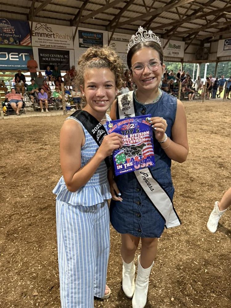 Two girls in pageant sashes and boots holding a booklet inside a livestock arena