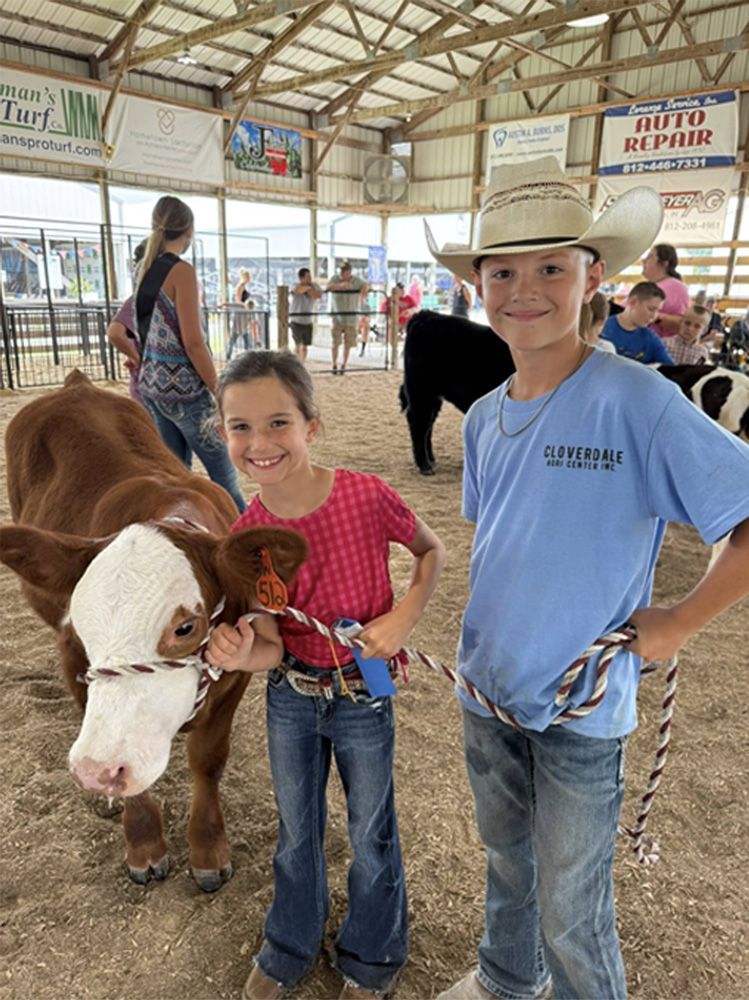Children stand with a cow at a livestock show inside a barn, one in a cowboy hat and blue shirt.