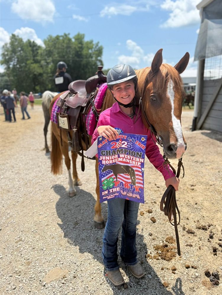 Child in a helmet holds a horse's lead rope beside a saddled horse at an outdoor riding area.