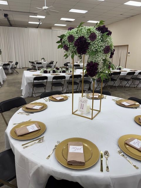 Round banquet table with gold plates, white napkins, and a floral centerpiece in a decorated event hall