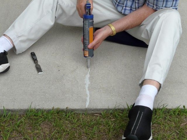 Person filling a crack in concrete with a sealant gun, seated outdoors, with a trowel nearby.