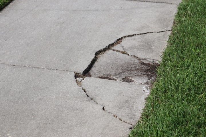 Damaged concrete sidewalk with large cracks, adjacent to green grass.