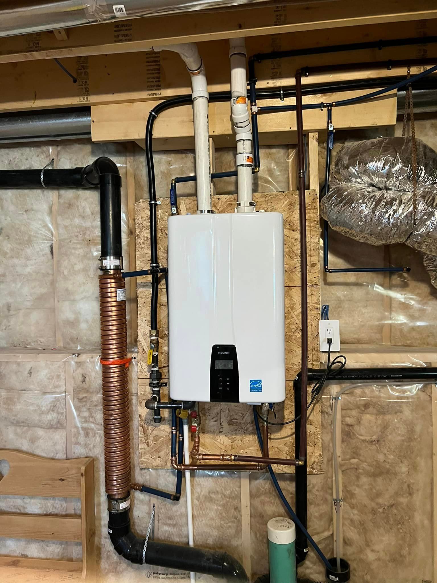 Tankless water heater mounted on a wall with various pipes, exhaust, and electrical components in a basement setting.