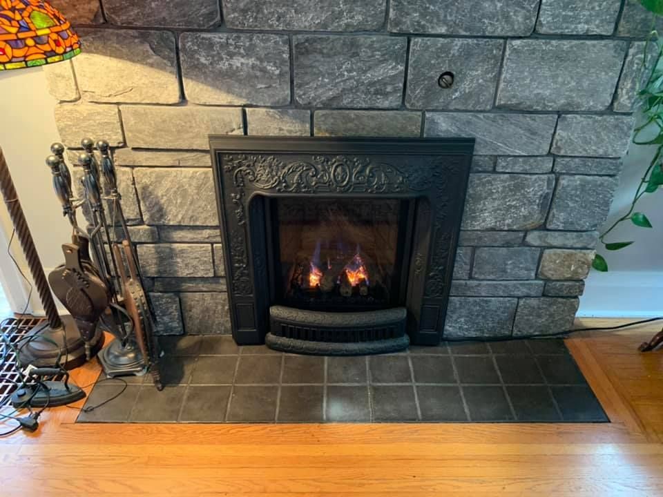 Fireplace with lit flames, dark tile hearth, stone surround, and ornate black metal insert. Fire tools to the left.