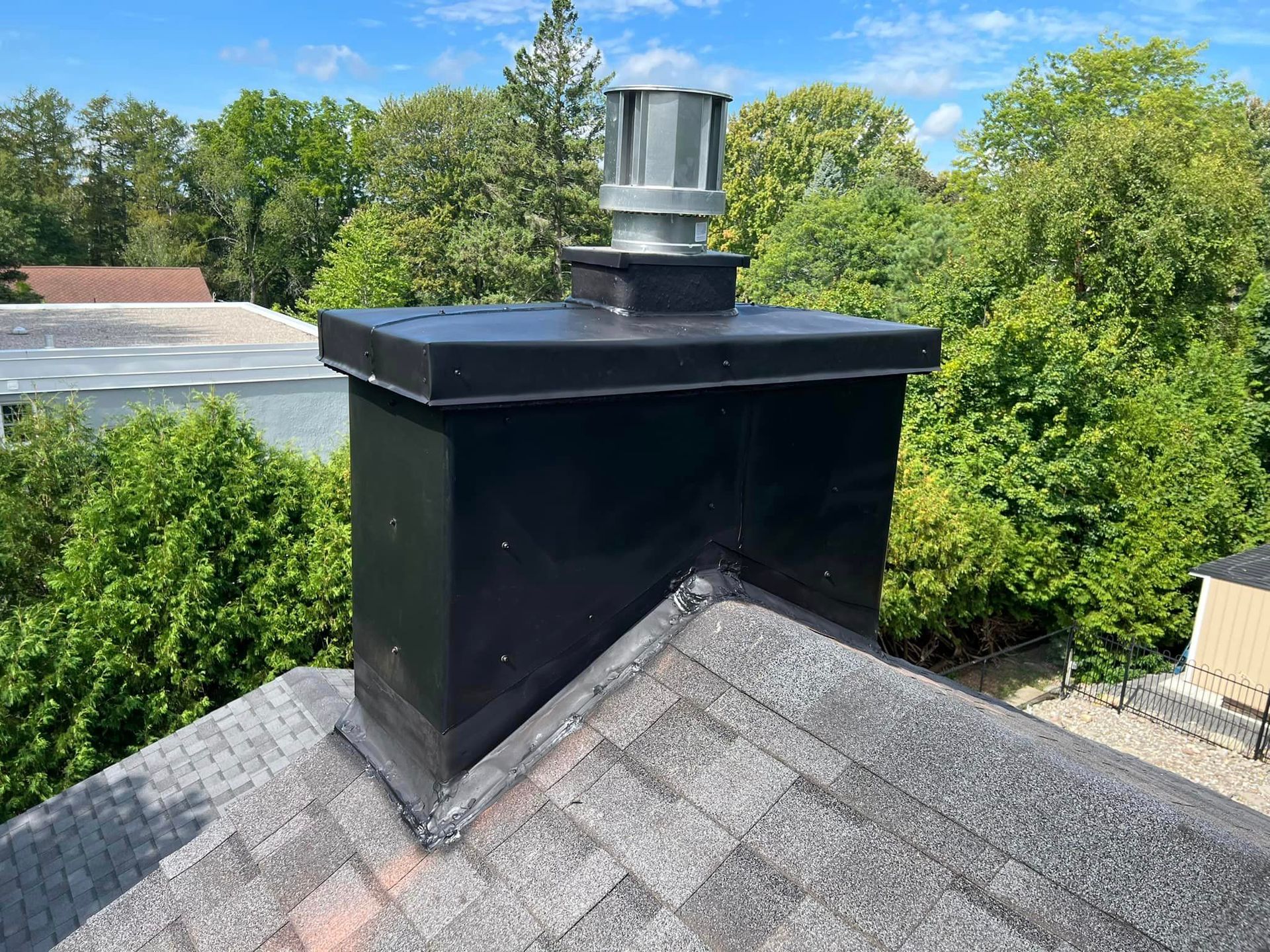 Black chimney with metal cap on a shingled roof, surrounded by trees.