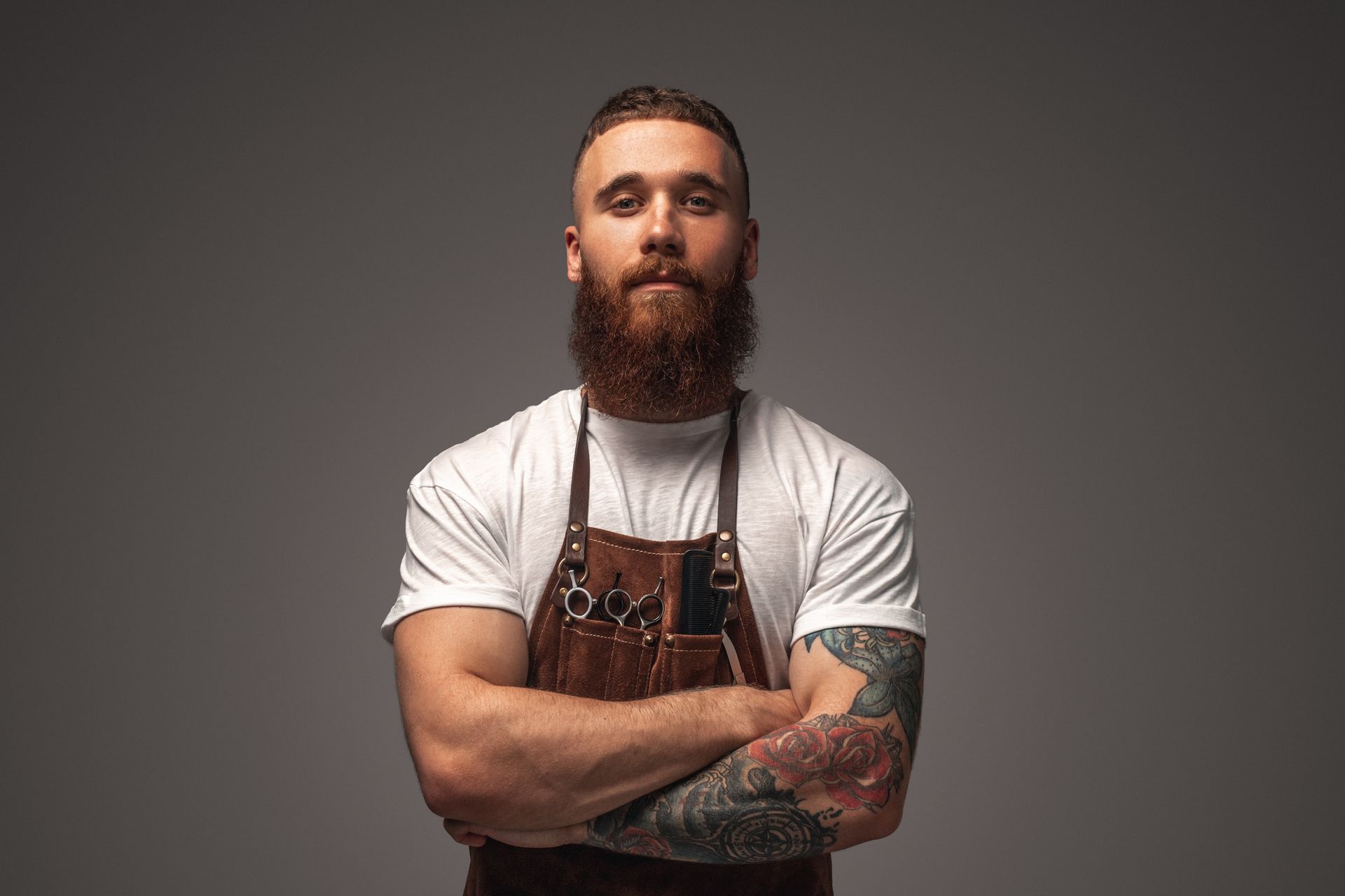 Man with full beard, arms crossed, wearing a leather apron and white t-shirt, standing against a gray backdrop.