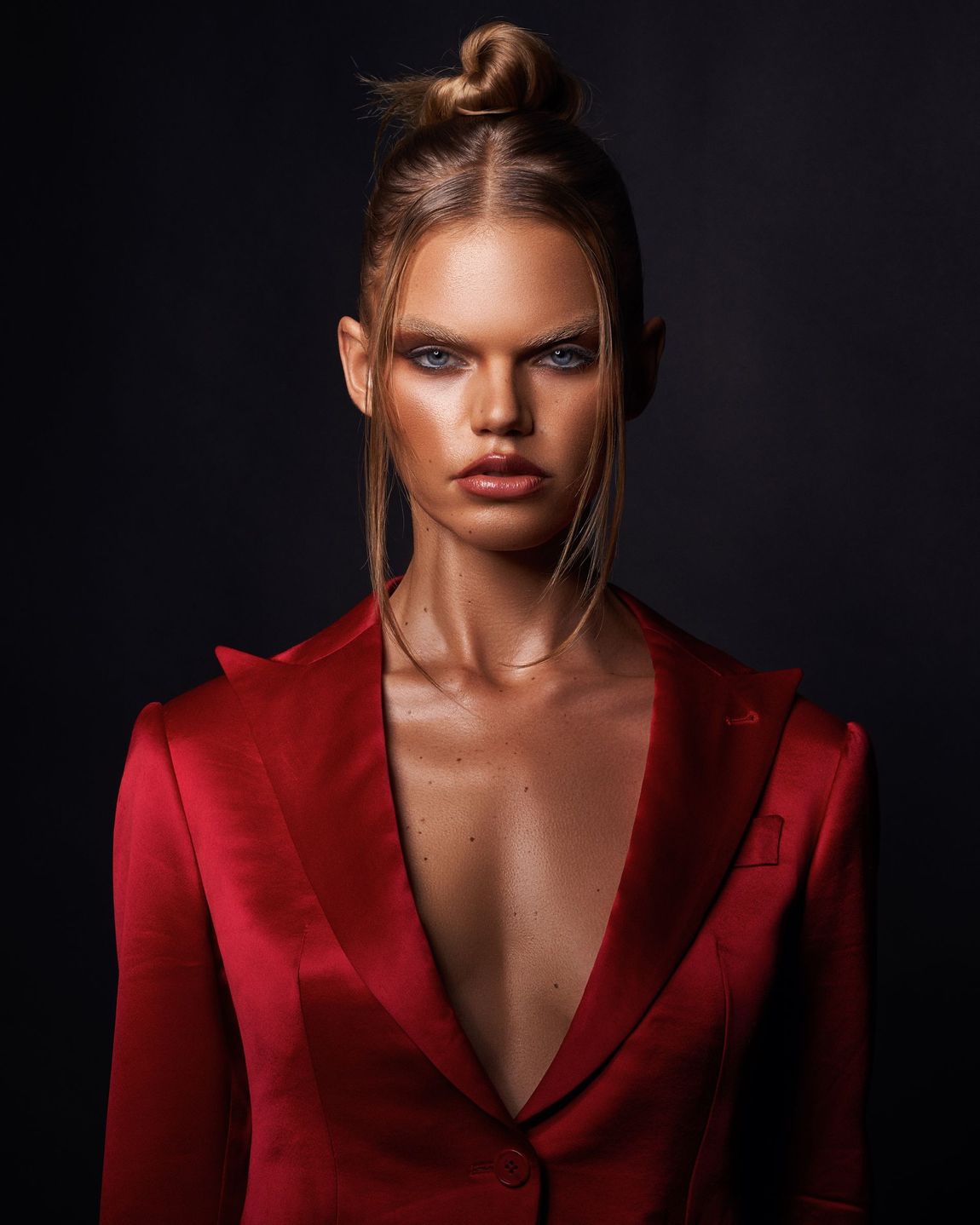 Woman in red blazer with bun, dramatic eye makeup, dark background.