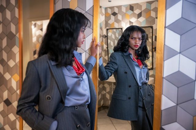 Woman in a blue pinstripe suit, looking in a mirror. Red necklace, blue blouse. Modern setting.