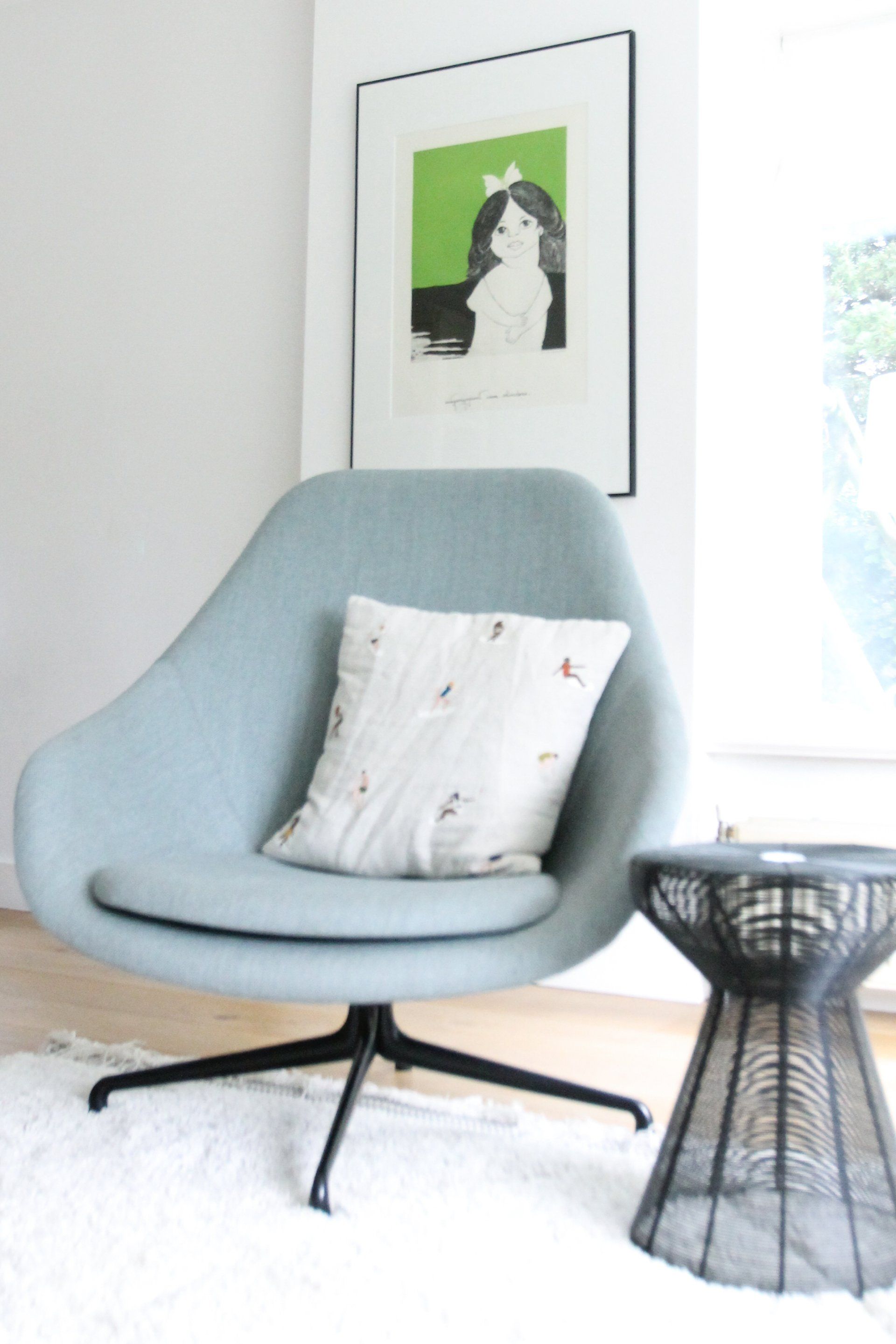 Lichtblauwe fauteuil met kussen, kunstwerk en zwart bijzettafeltje in een lichte kamer met een wit vloerkleed.