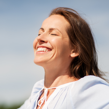 Smiling person outdoors in white clothing, eyes closed in bright sunlight