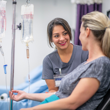 Nurse smiles while talking with a patient in a hospital bed beside IV drip bags.