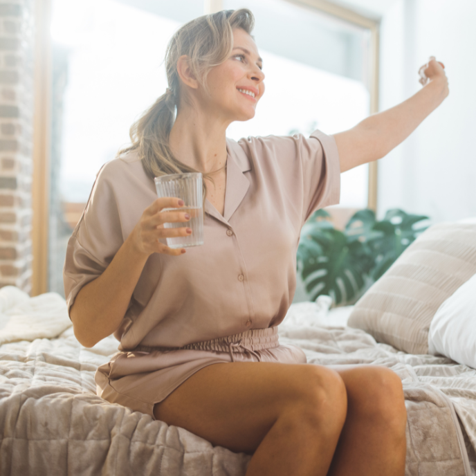 Person sitting on a bed, smiling and raising a glass in a bright bedroom
