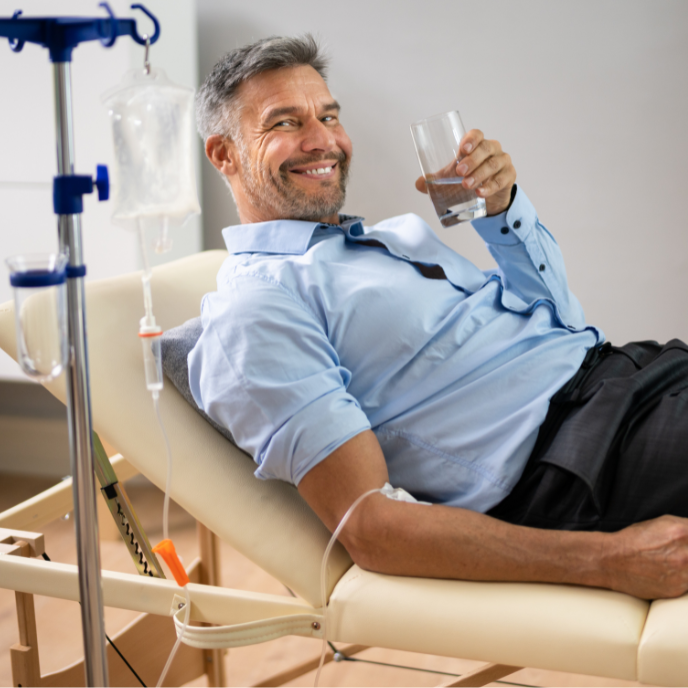 Relaxing patient reclining in a chair, smiling and holding a glass beside an IV drip stand.