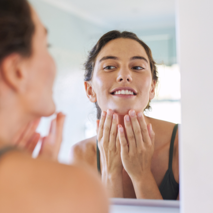 Woman examining her face in a mirror, touching her chin in a bright bathroom.
