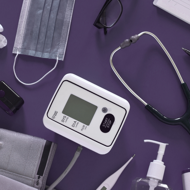 Blood pressure monitor, stethoscope, face mask, glasses, and sanitizer on a purple background.