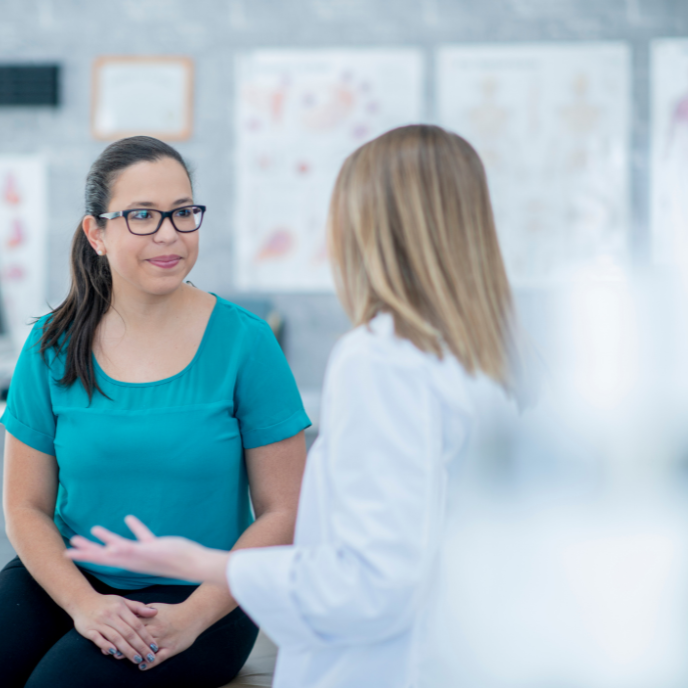 Two women talking in a clinic, one seated in a teal shirt and one in a white coat.