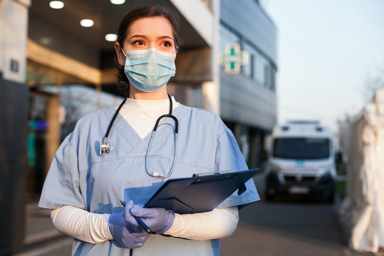 A nurse wearing a mask and holding a clipboard is standing in front of a hospital.