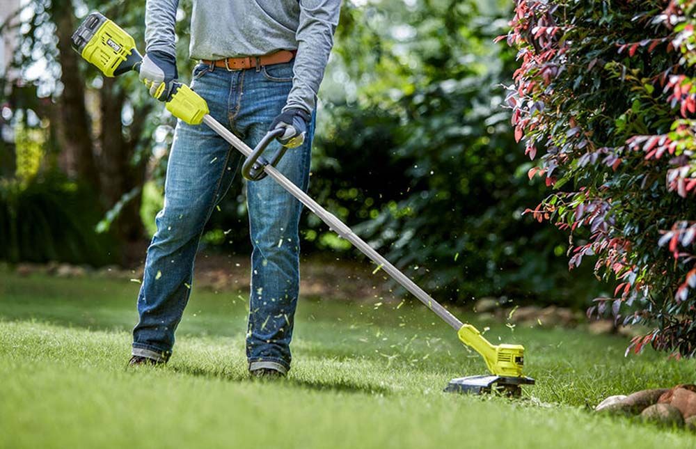 Person using a green and black string trimmer on a grassy lawn; debris flying.