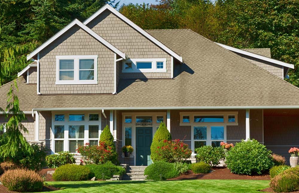 Two-story home with a brown roof, beige siding, and green lawn. Landscaped with shrubs and trees.