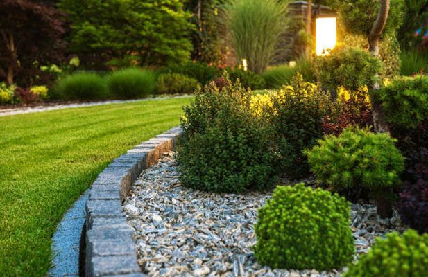 Lush garden with green lawn, stone border, gravel bed, and various shrubs and plants. Light post glows in the background.