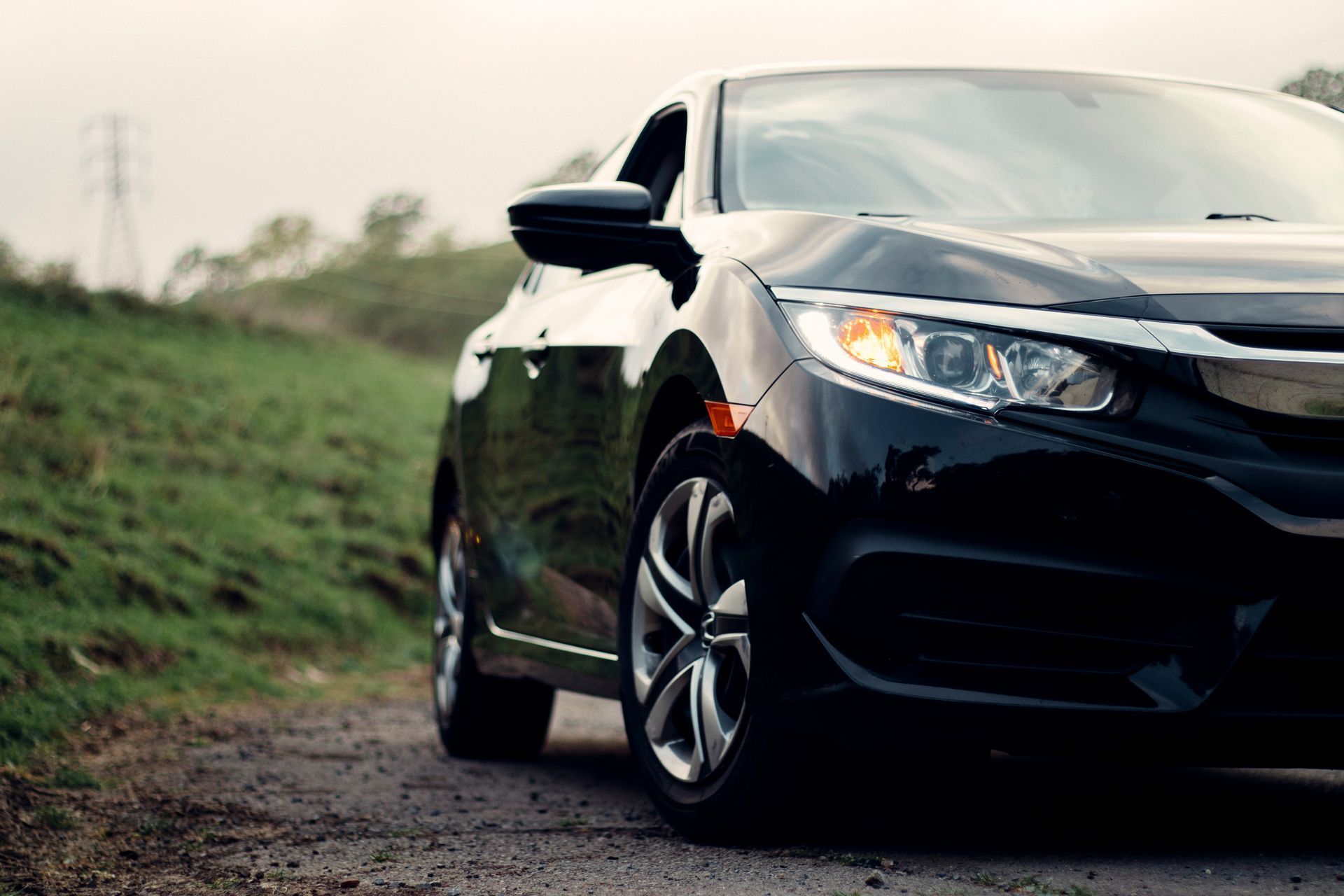 Black sedan parked on a gravel road beside a grassy field under an overcast sky