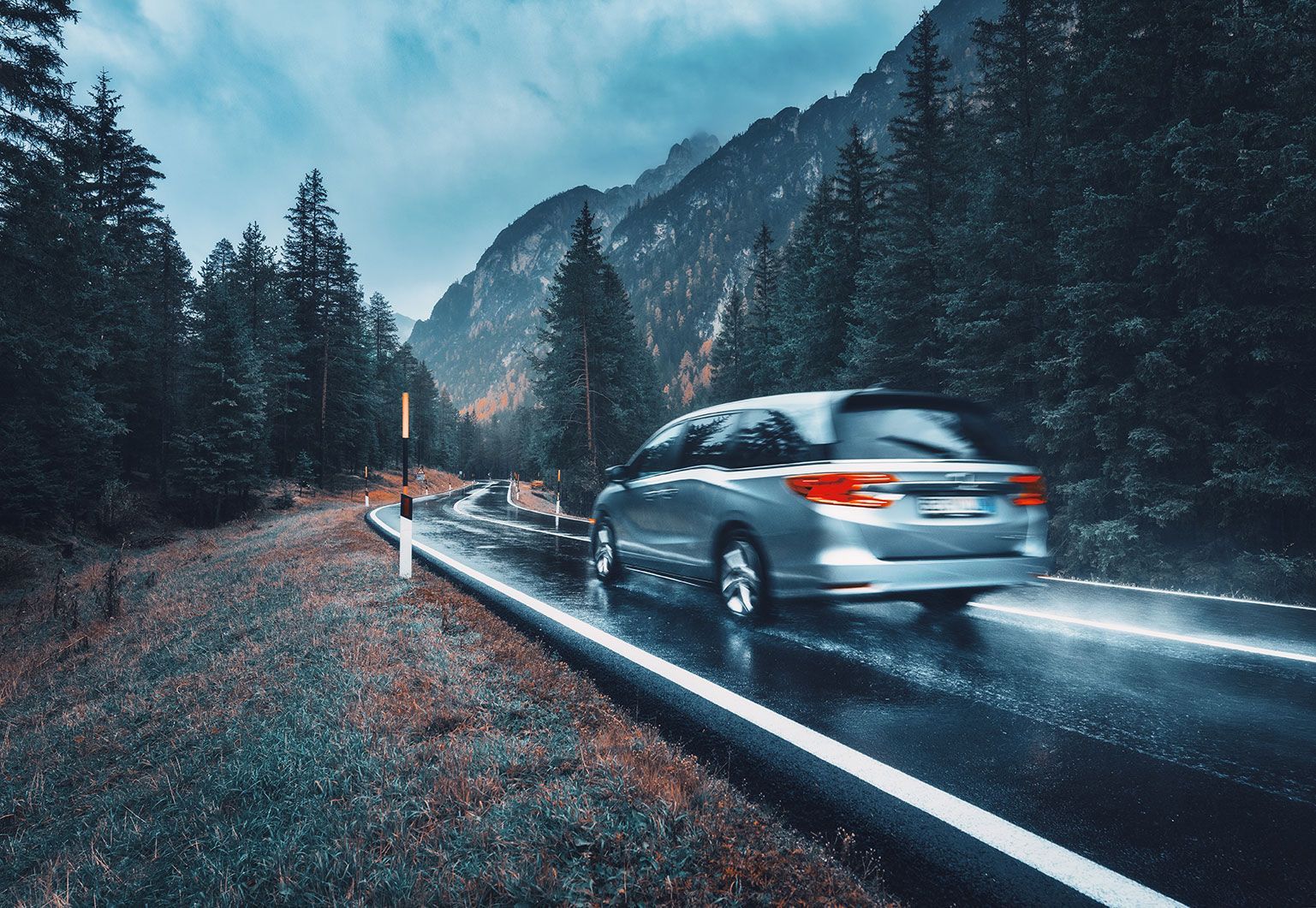 Silver SUV driving on a wet mountain road through a pine forest in the rain