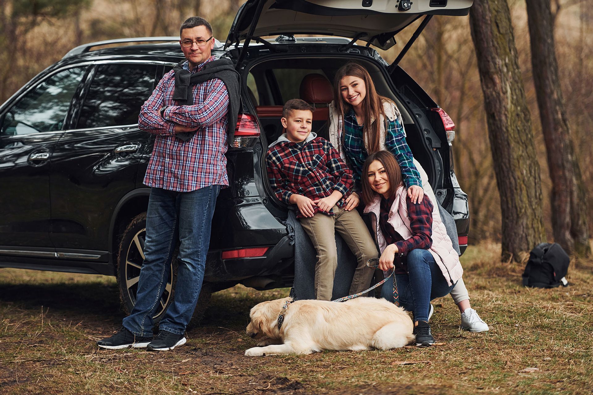 Family posing beside open SUV in a wooded area, with a light-colored dog lying in front.