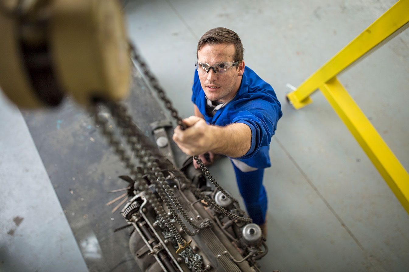 Man in blue workwear reaching up with a chain hoist near industrial machinery in a factory.