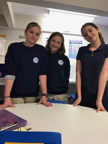 Three young girls are standing next to each other at a table in a classroom.