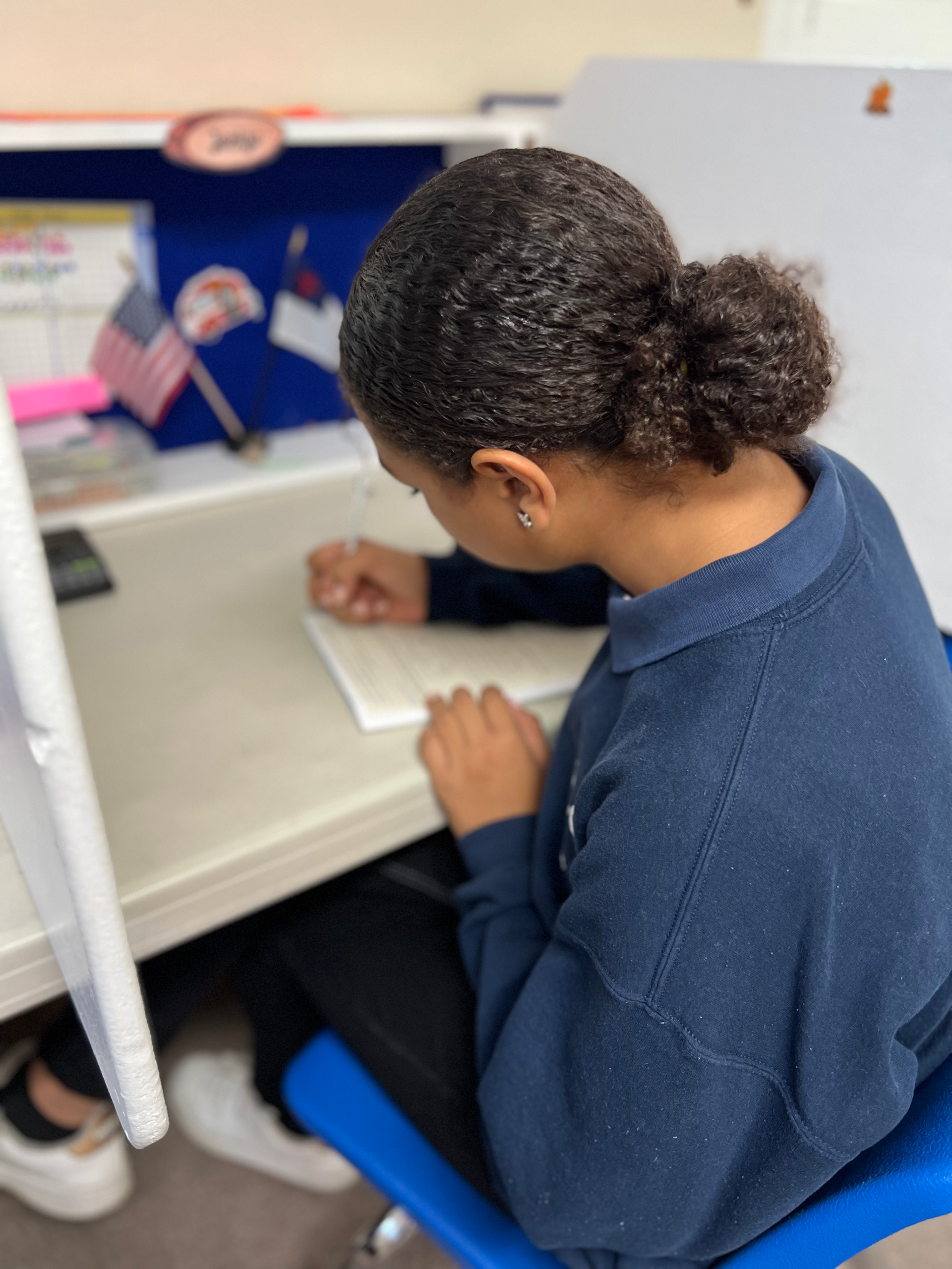 A girl in a blue shirt is sitting at a desk writing in a notebook