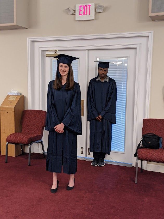 A woman in a graduation cap and gown stands in front of an exit sign