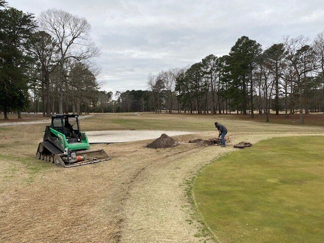 Skid steer leveling a dirt path beside a golf course green, with a worker nearby
