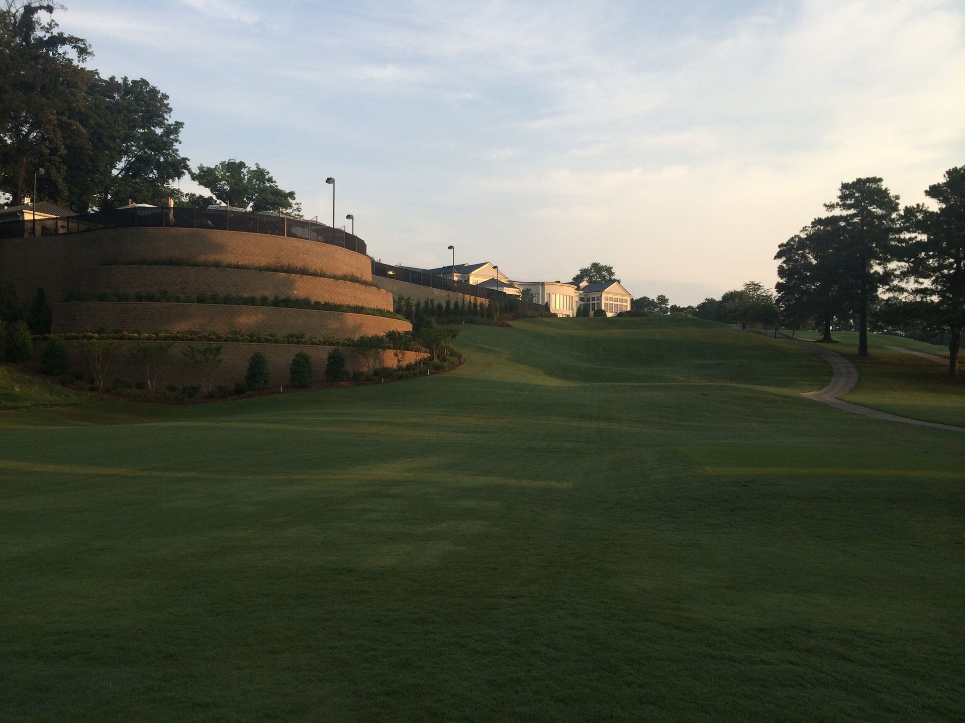 Golf course fairway beside a terraced hill at sunset, with trees and distant buildings.