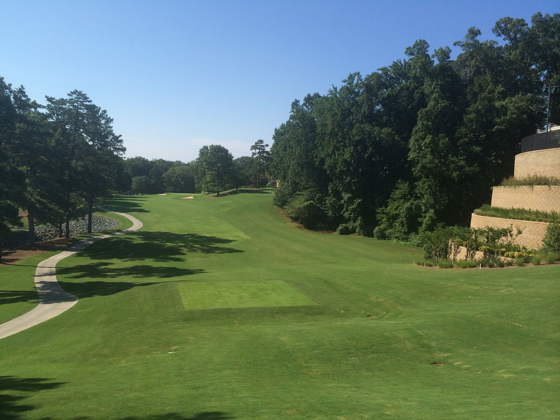 Sunny golf course fairway with green hills, trees, and stone retaining wall along the right side