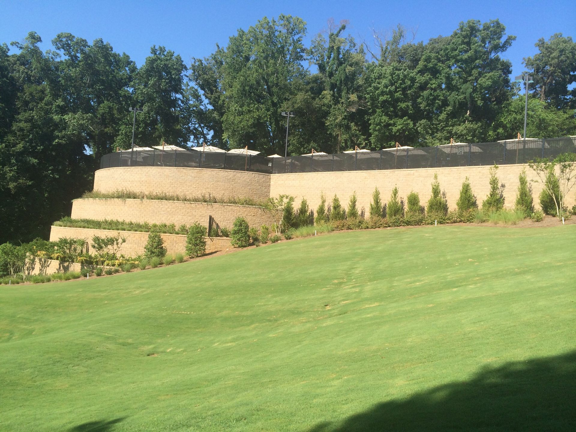 Manicured green lawn below terraced tan retaining walls and trees under a clear blue sky
