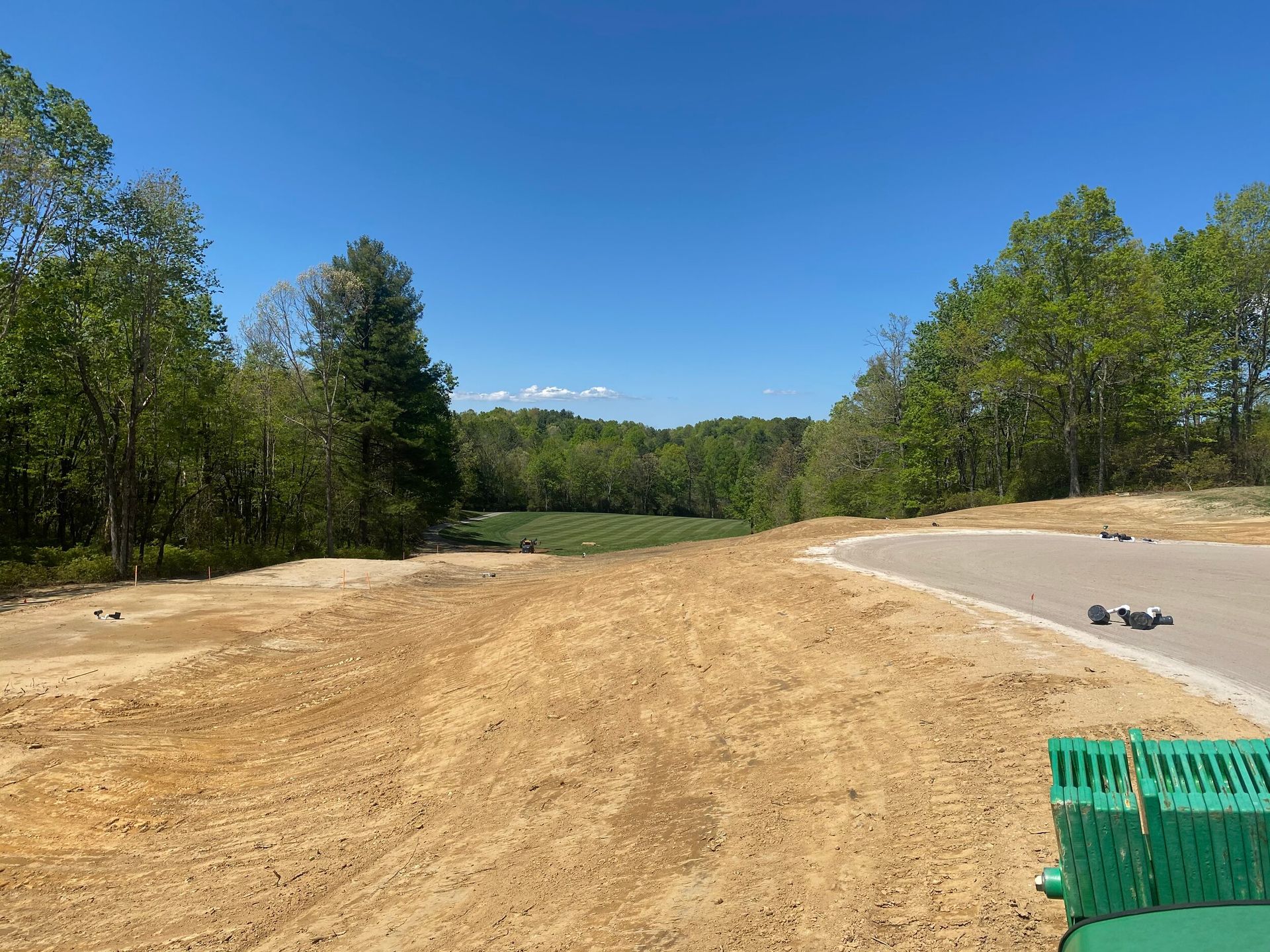Dirt construction site beside a paved road, with trees and a clear blue sky