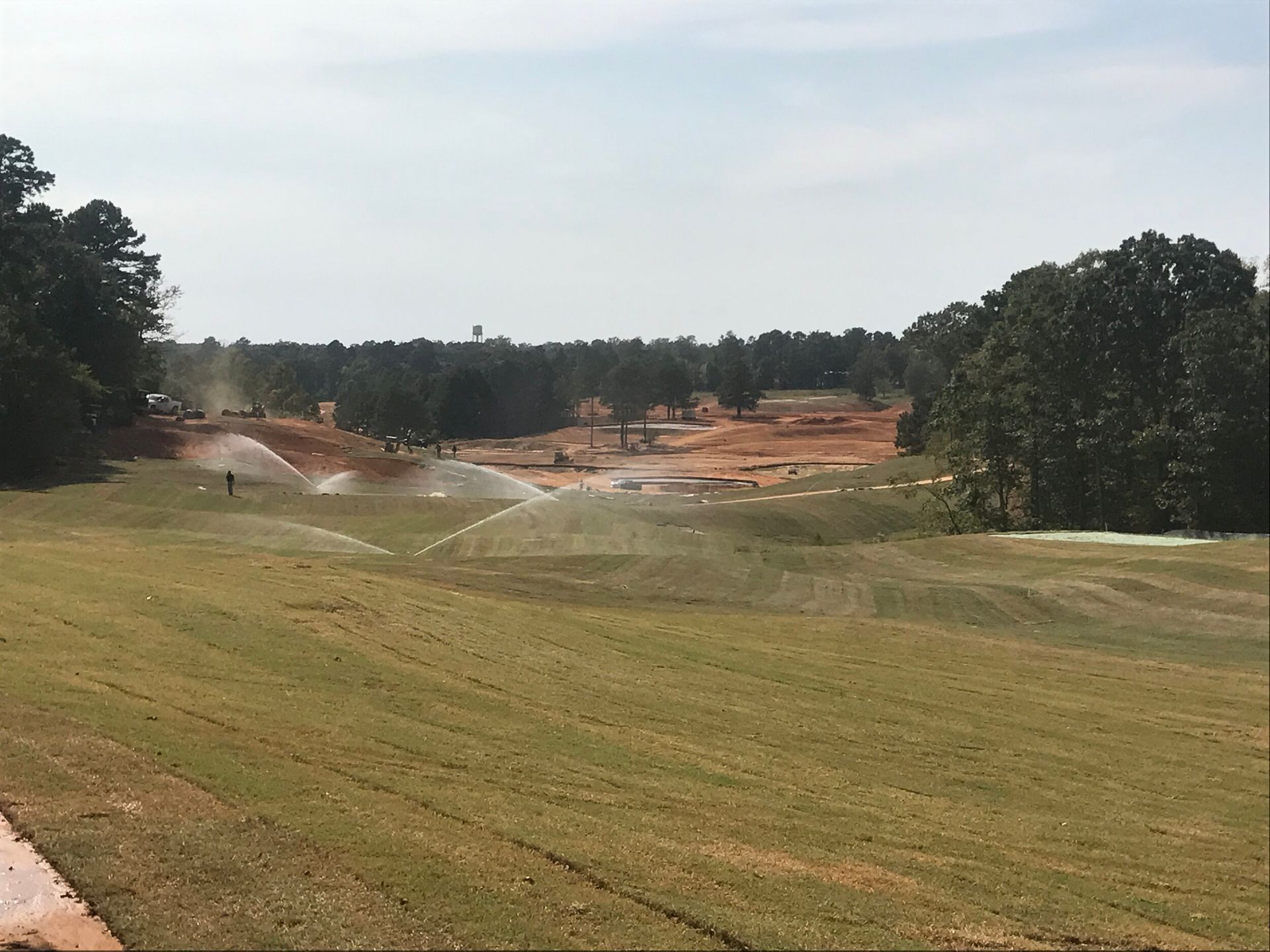 Grassy field with sprinklers spraying water near a wooded, orange dirt construction area under a cloudy sky