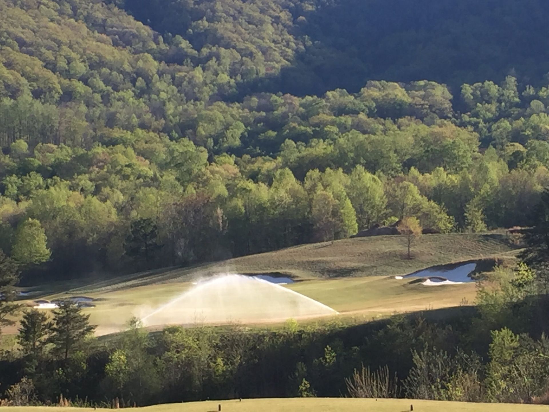 Sprinklers watering a grassy field beside a wooded hillside on a sunny day