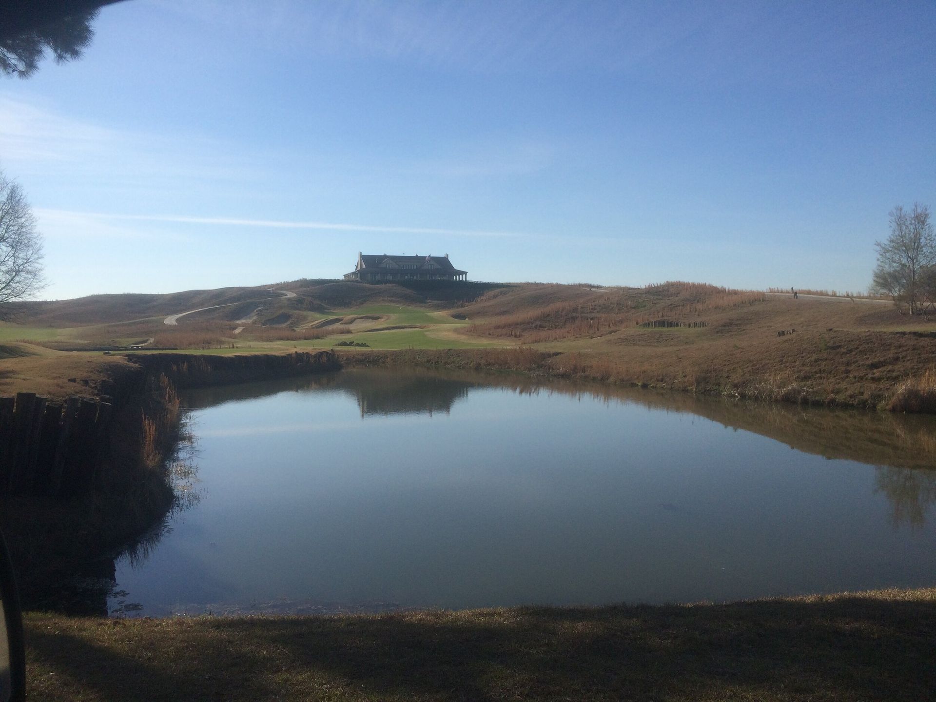 Calm pond reflecting blue sky, with grassy banks and a distant building on the horizon.