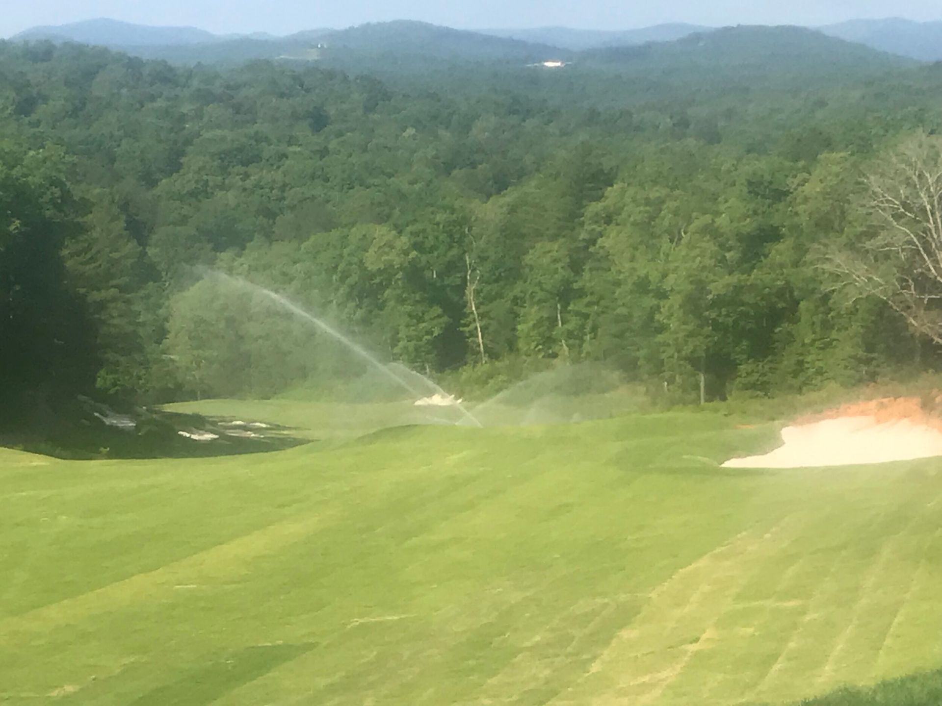 Wide grassy golf course with sprinklers spraying water, backed by dense green forest and distant hills