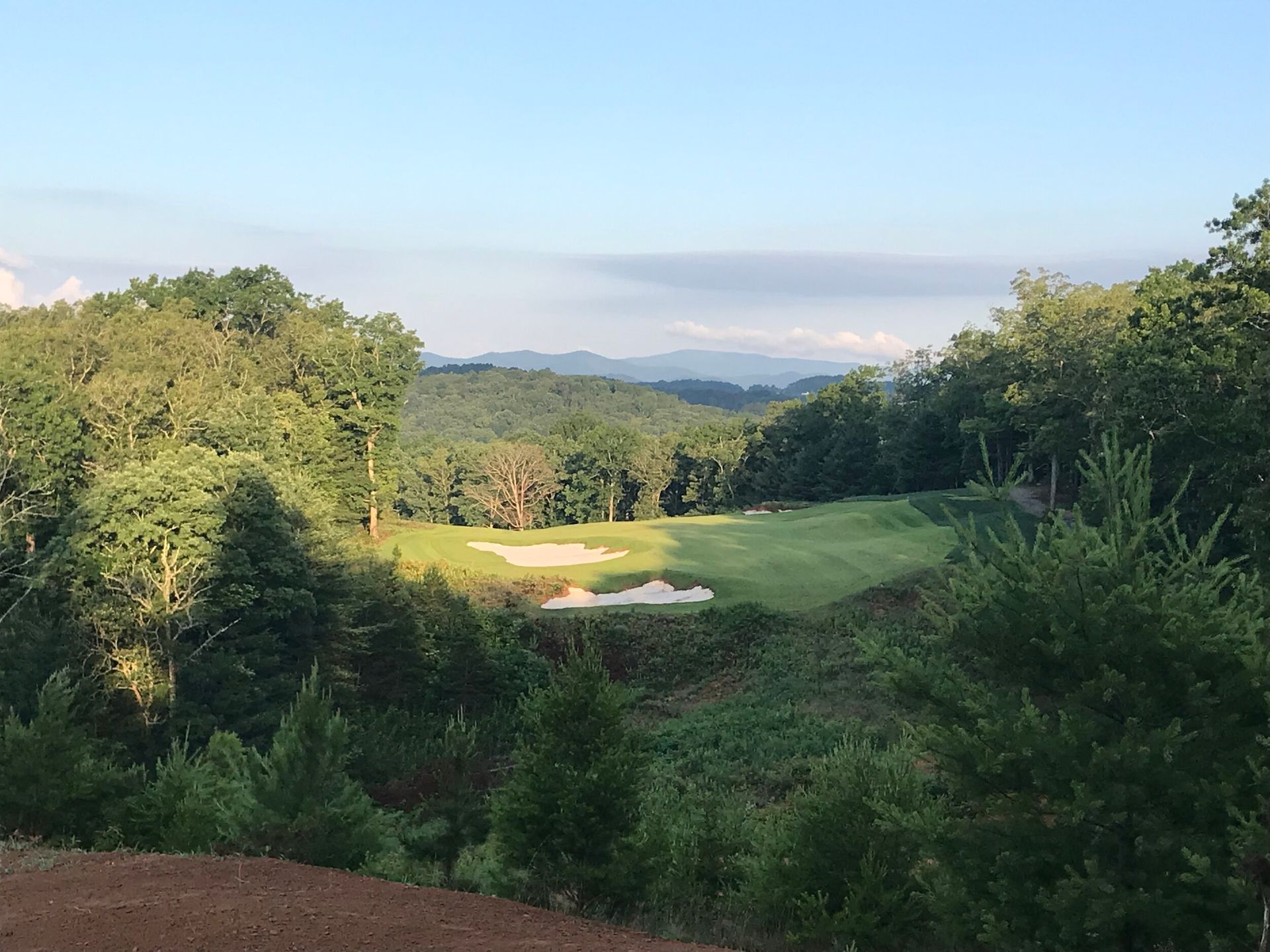 Sunlit golf course green framed by trees, with distant hills under a clear sky