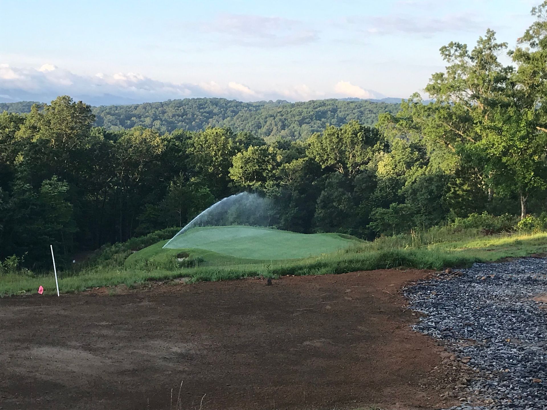 Grassy hill with a sprinkler spraying water, bordered by trees and distant mountains under a clear sky