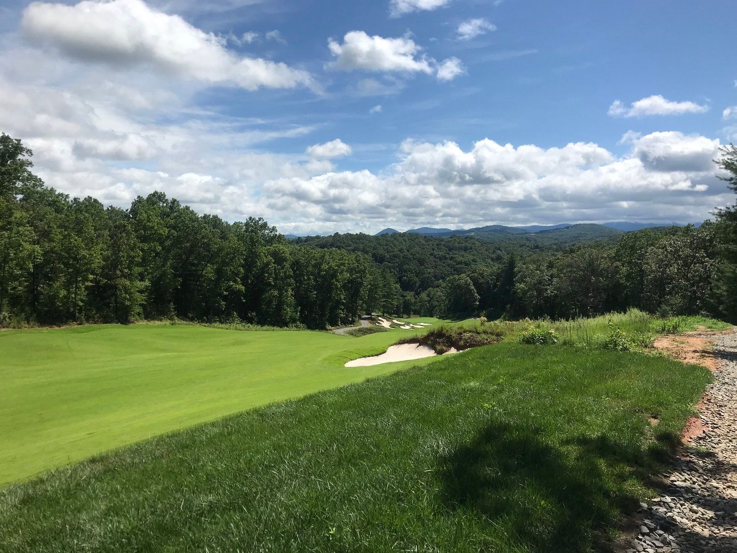 Lush golf course fairway with sand bunker, trees, and a partly cloudy sky over distant hills