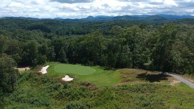 Green fairway overlooking forested hills and cloudy mountains, with sand bunkers and a winding path