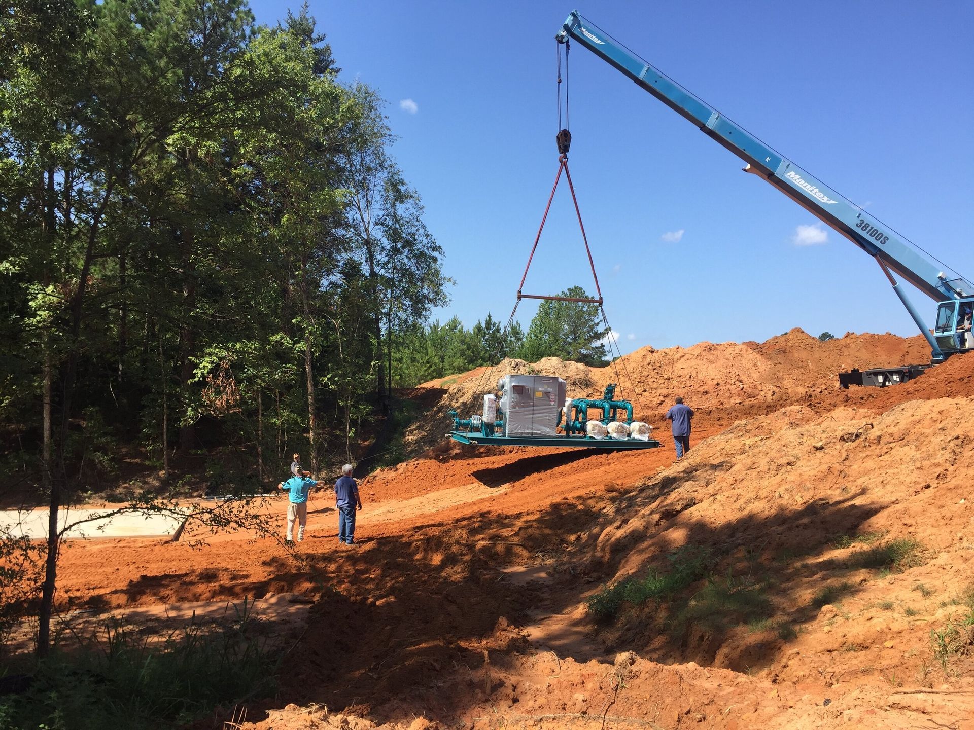 Construction site with crane lifting a load over a red dirt hillside, workers nearby under blue sky