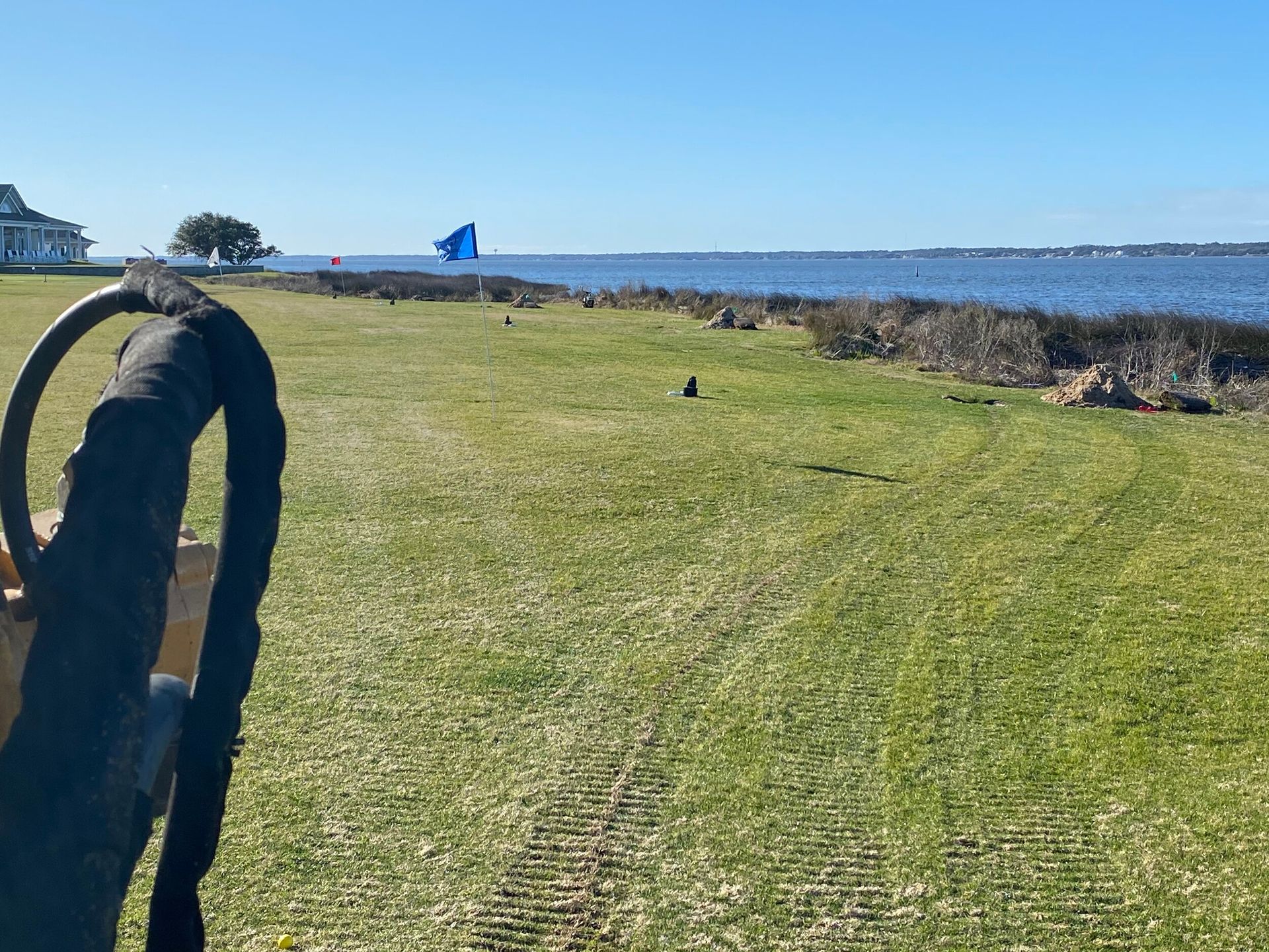 Grassy waterfront path with a railing, blue water, and a small blue flag in the distance